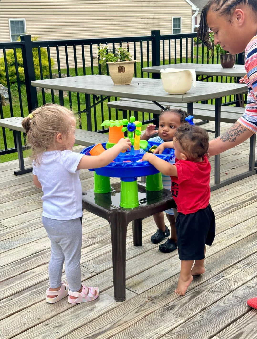 Three toddlers playing with a water table on a wooden deck, supervised by an adult.