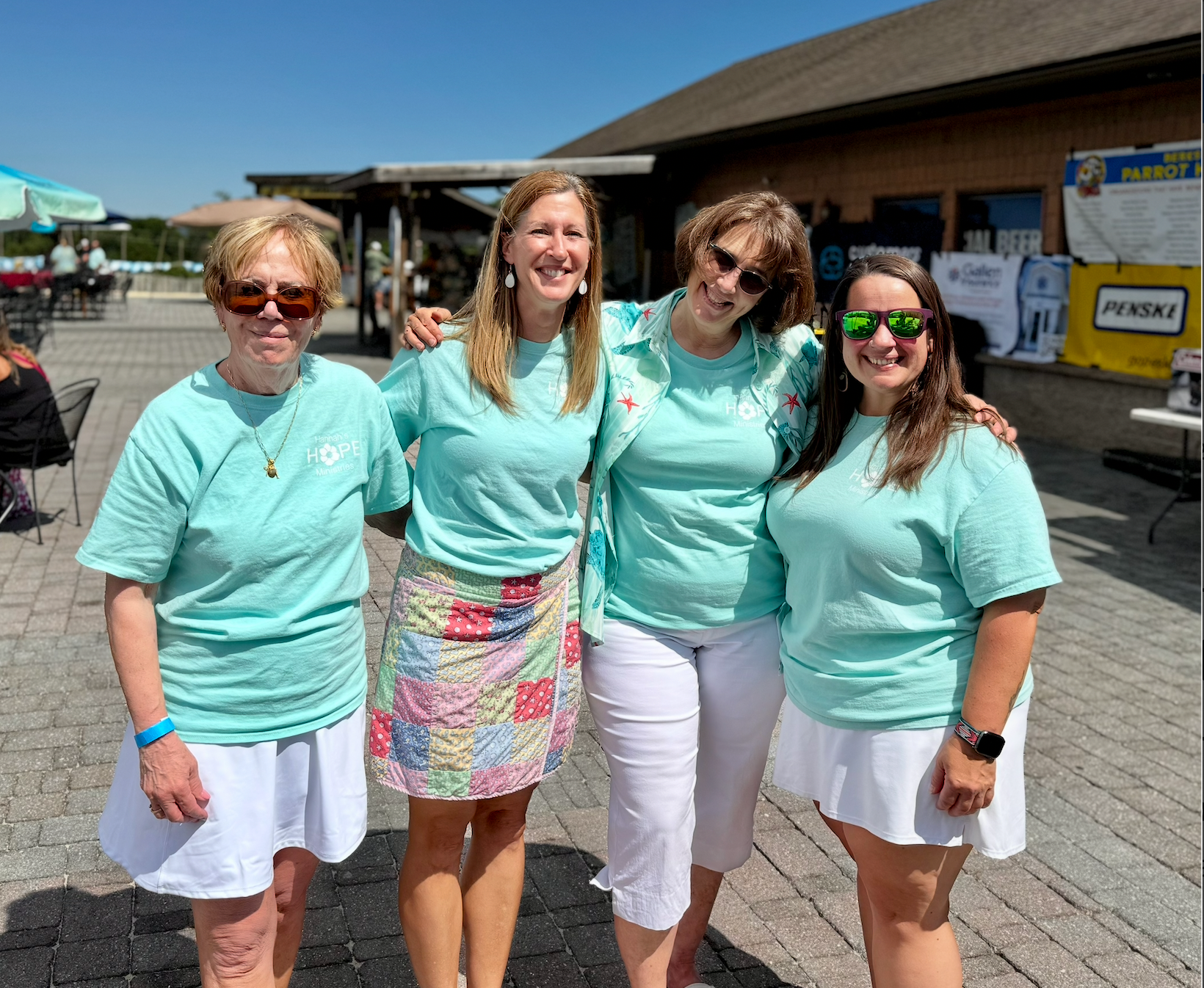 Four people smiling, posing outdoors. All wearing teal shirts; two in white skirts, one in a colorful skirt, one in white pants.
