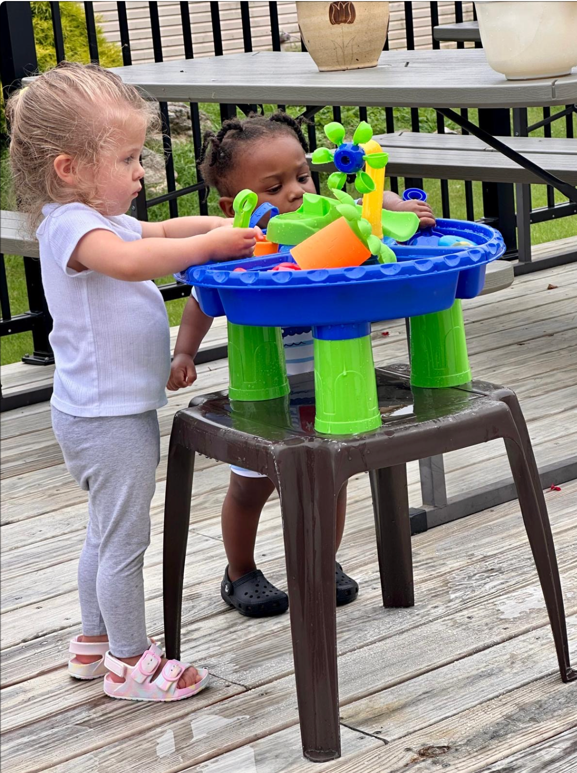 Two children playing with a water table on a deck; one is pouring water while the other watches.