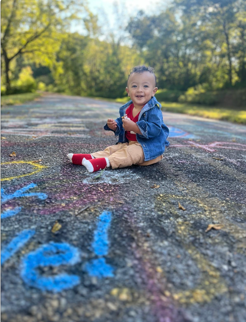 Young child sits on a chalk-covered path, wearing a denim jacket, red shirt, and beige pants, smiles at the camera.