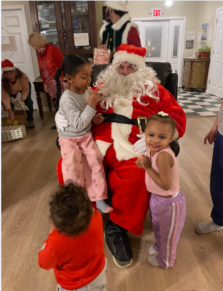 Santa Claus sits with children indoors. Several kids gather around.