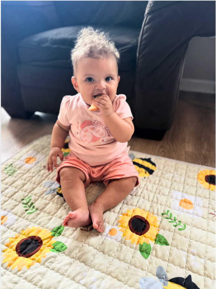 Baby sitting on a quilt, eating a snack. Wearing pink outfit.
