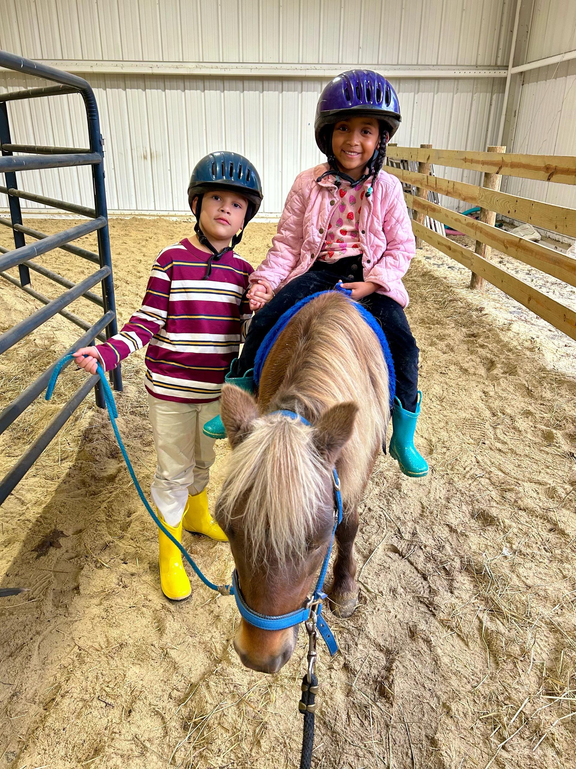 Two children wearing helmets; one on a pony, the other holding a lead rope in a riding arena.