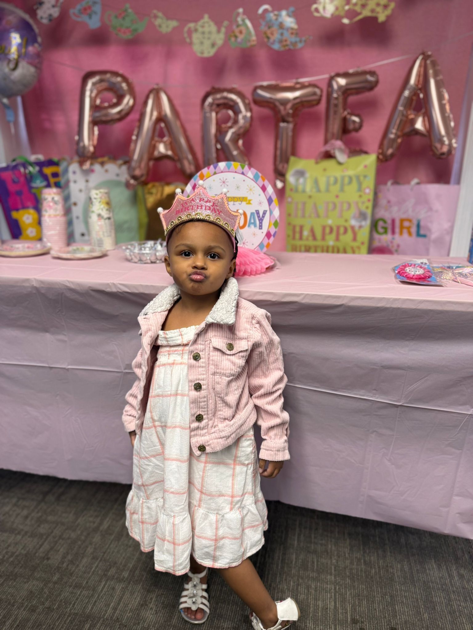 Young child at a party; wearing a pink jacket and dress, standing in front of party decorations, making a face.