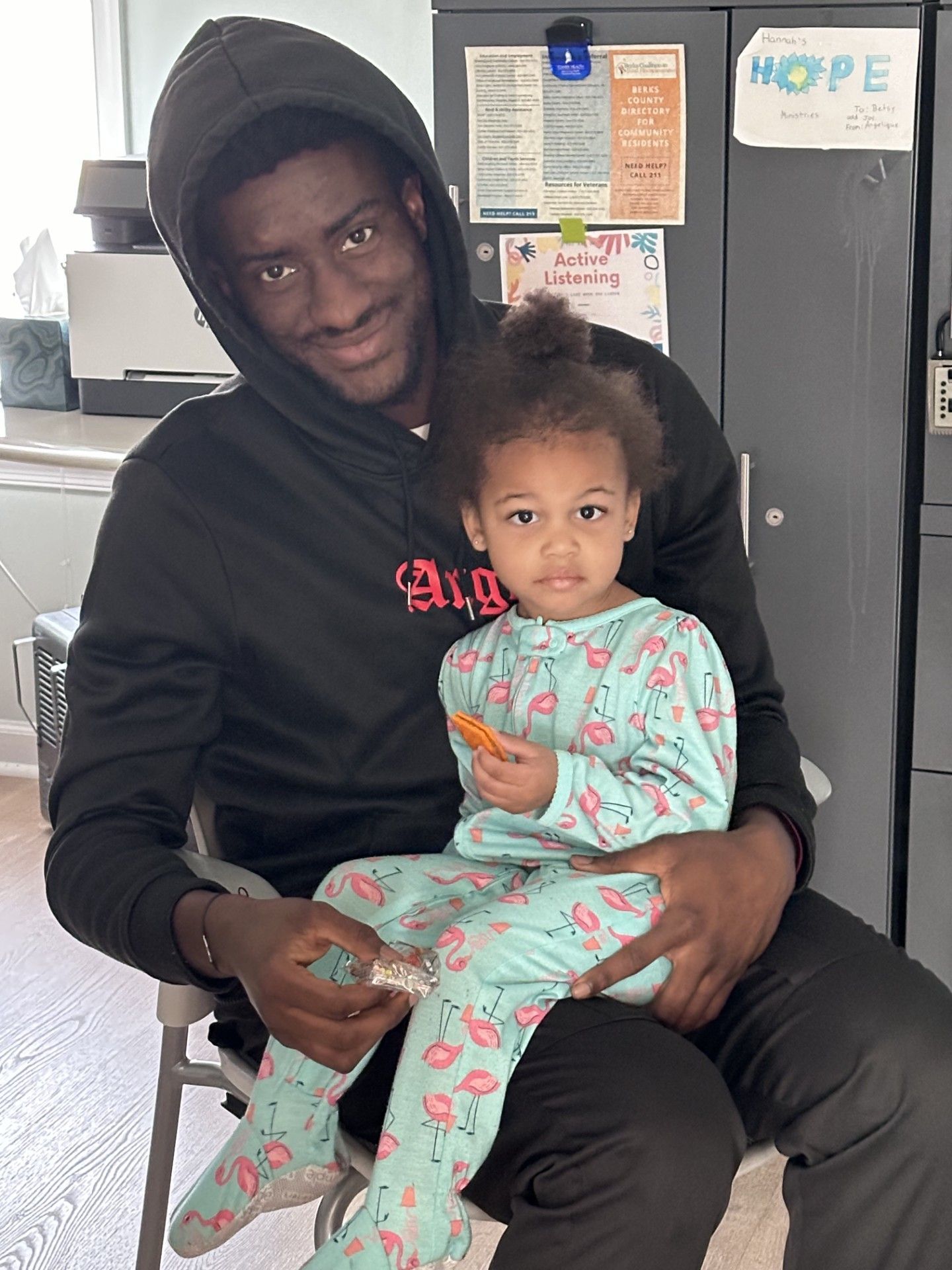 Man in black hoodie smiles, holding a child in flamingo pajamas. They sit indoors near a cabinet.