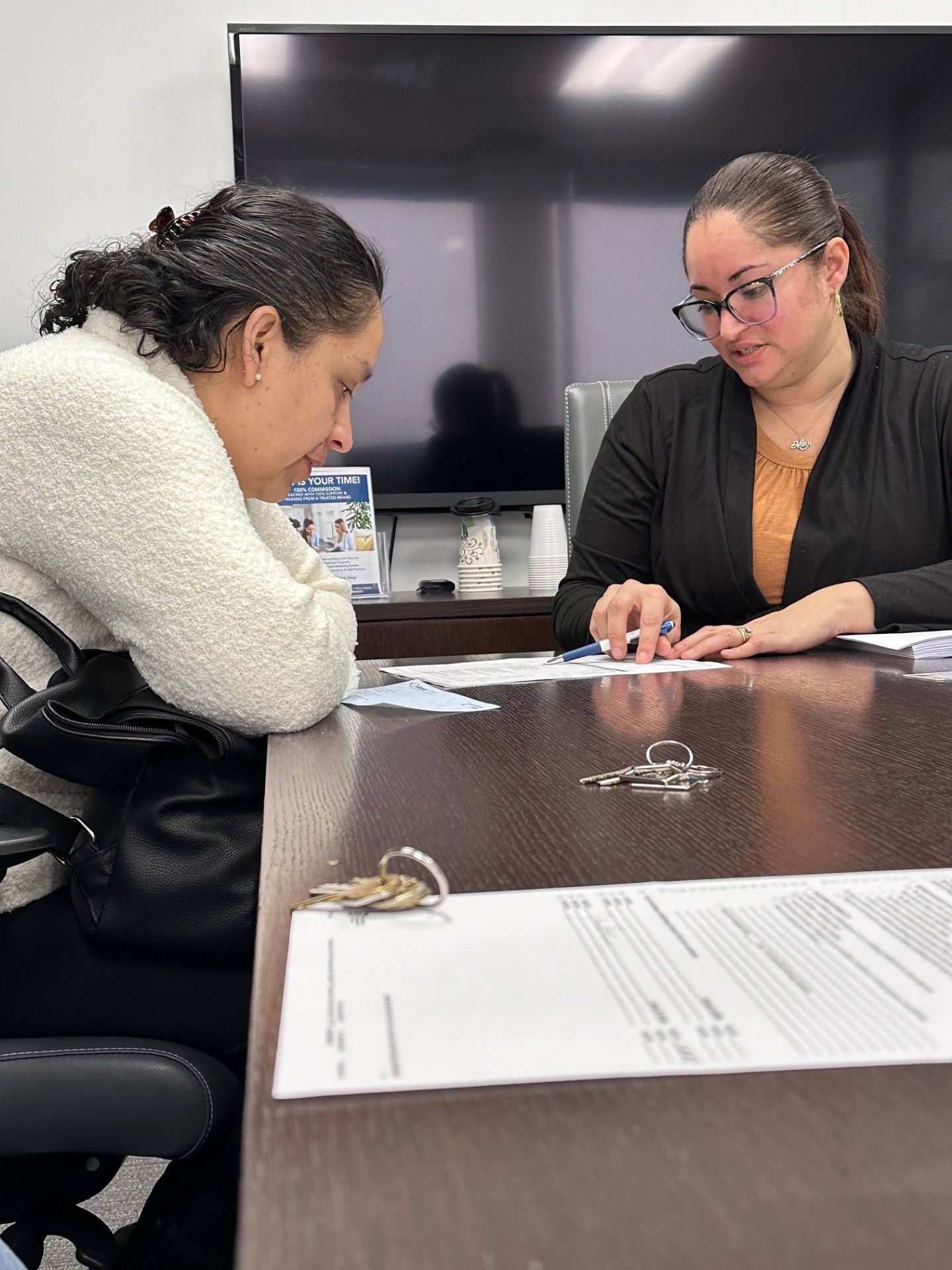 Two women at a wooden table, looking at a laptop and documents. One points. Bright room.