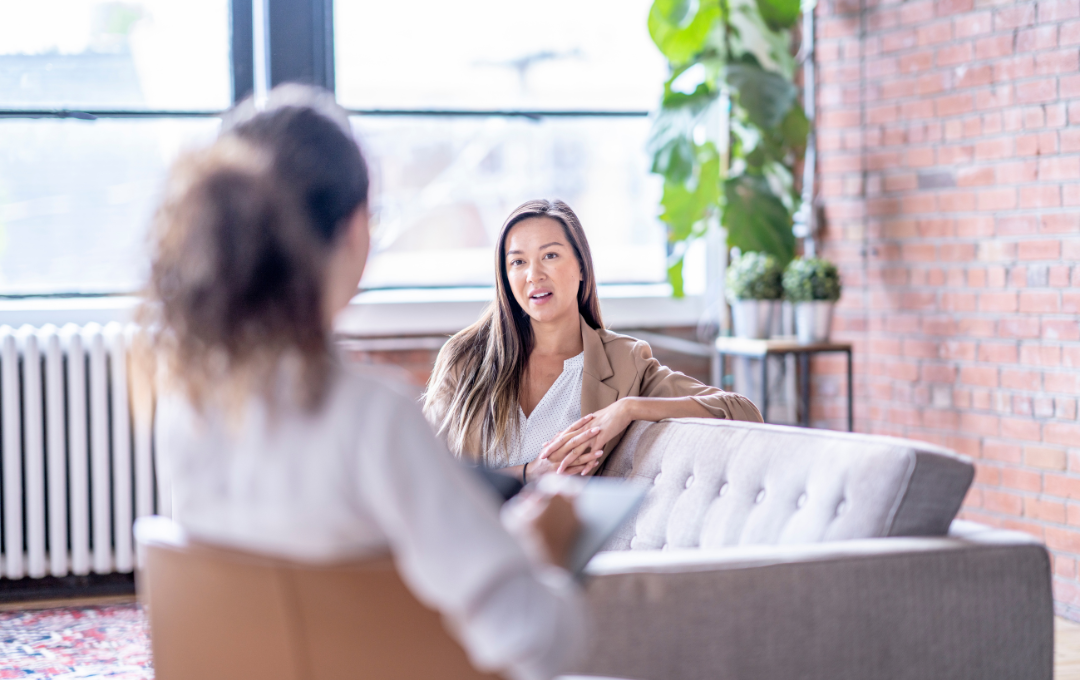 Woman on couch talking to another woman in a light-filled room with exposed brick and a plant.