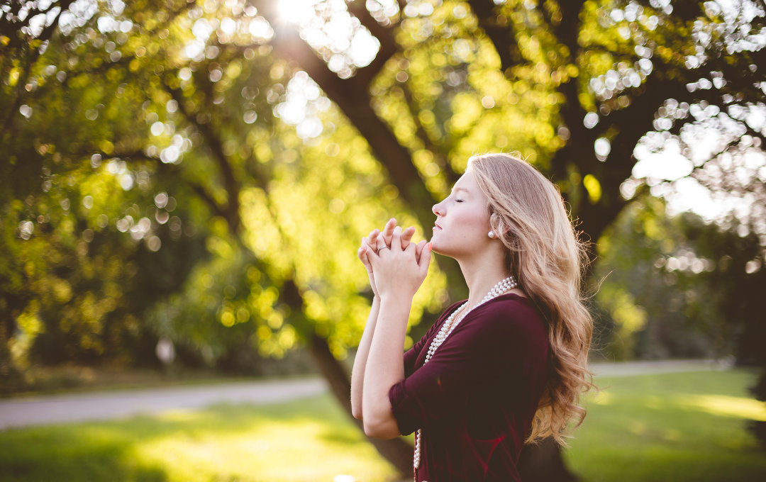 Woman with hands clasped, eyes closed, standing in a sunny park setting, trees in the background.