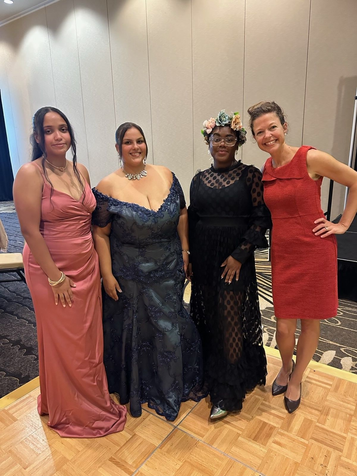 Four women in formal attire pose indoors; the woman on the left in a pink gown, another in a blue gown, and one in a black gown, the fourth in a red dress.