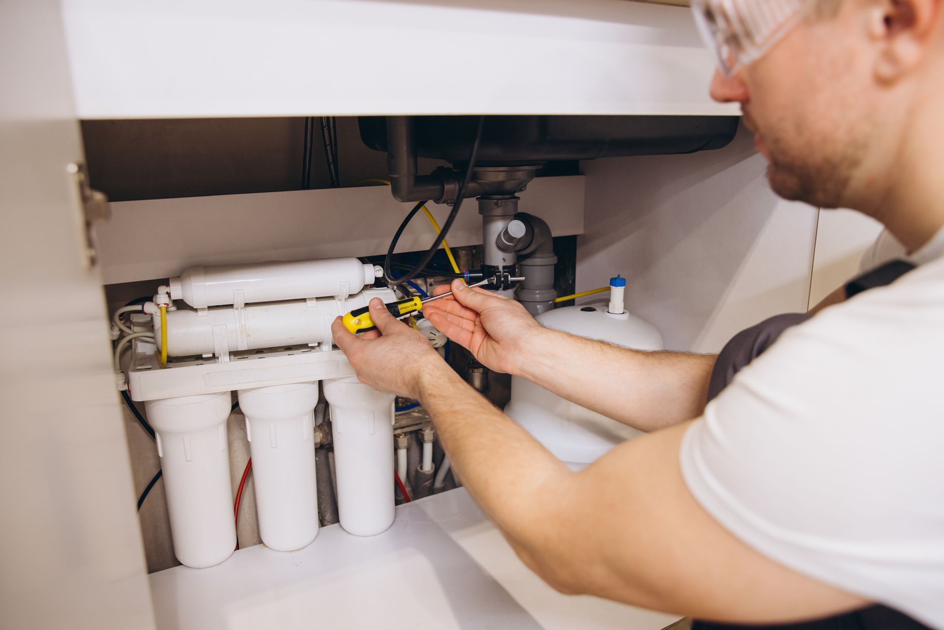A person wearing safety glasses repairs a water filter system under a sink.
