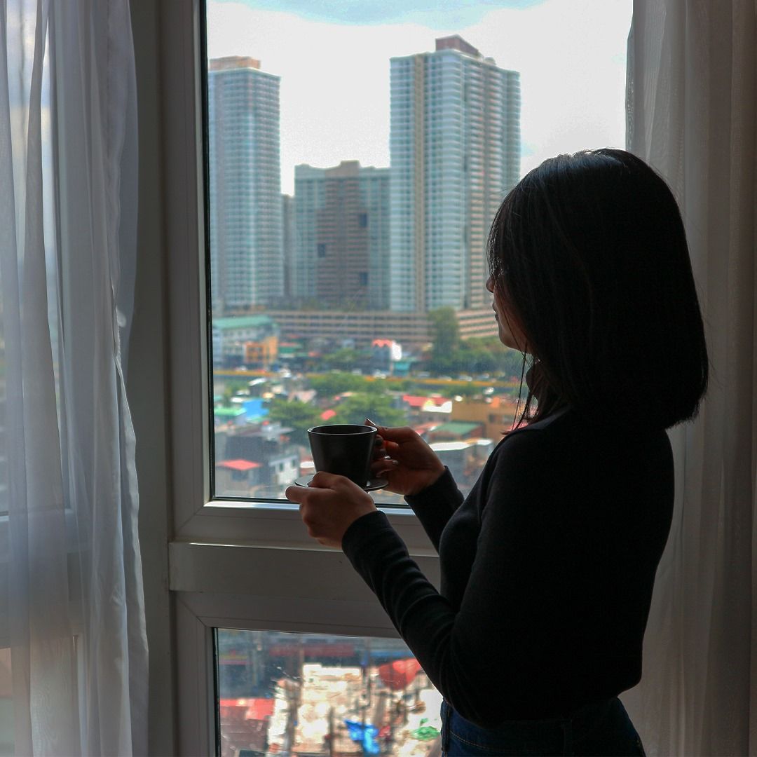 Silhouetted person holding a cup by a window, looking out at tall city buildings.