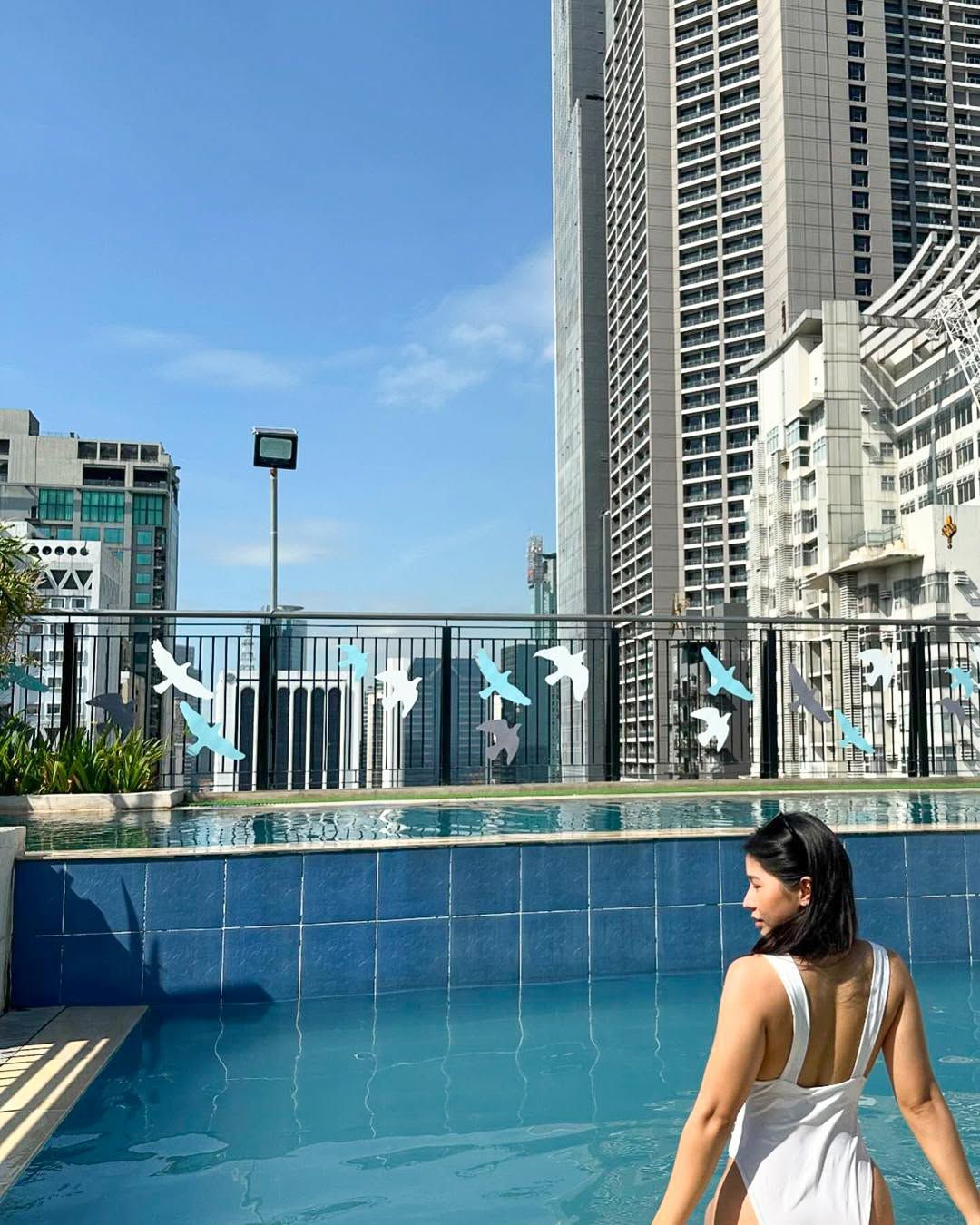 Woman in a white swimsuit by a rooftop pool, city skyline background, blue water, sunny day.