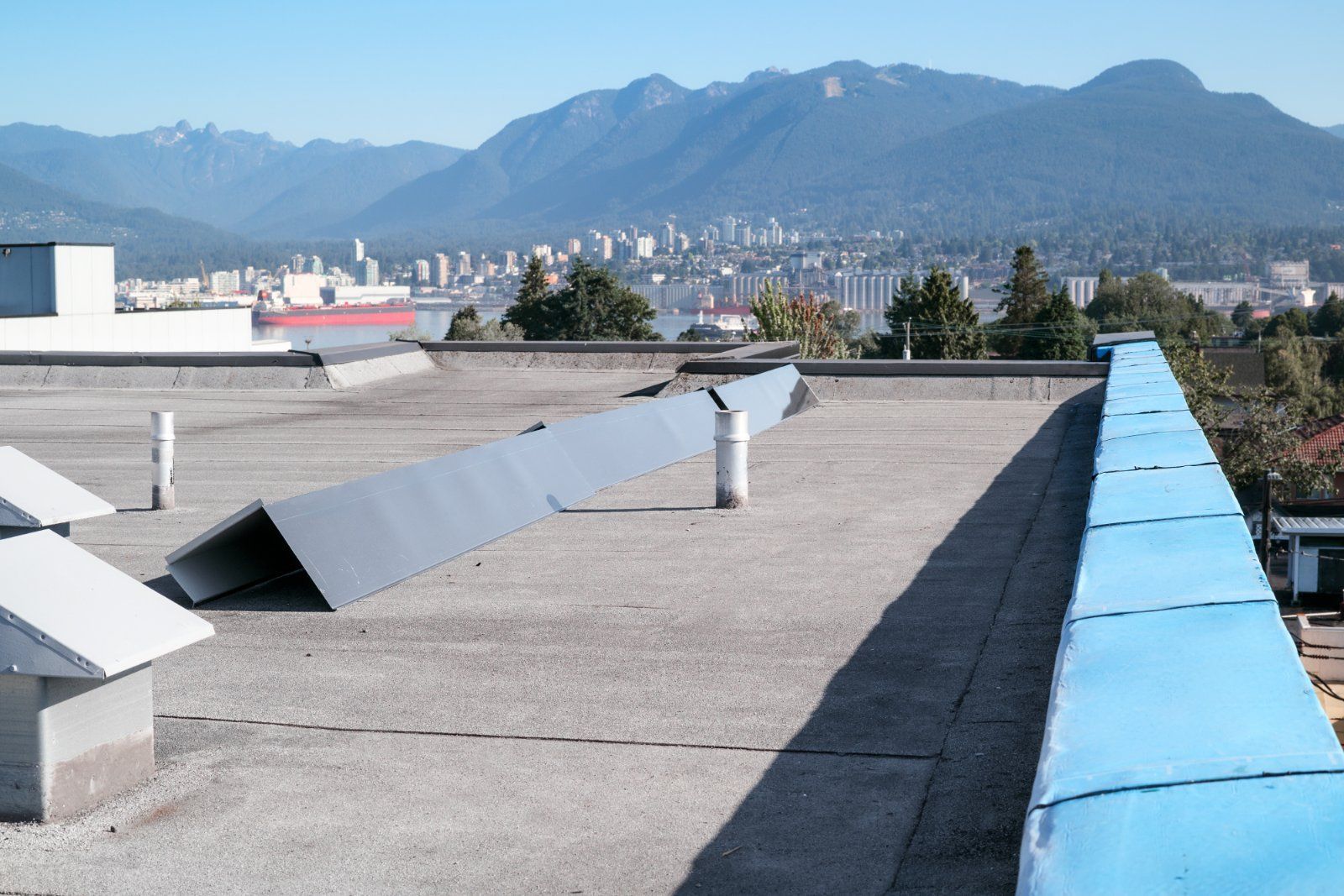 Flat rooftop with metal vents and a blue-topped parapet, overlooking a distant harbor and mountains under a blue sky.