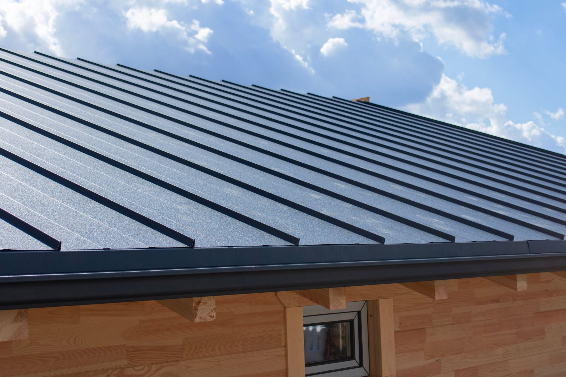 A dark grey standing-seam metal roof on a light-colored wooden building under a blue sky with scattered clouds.