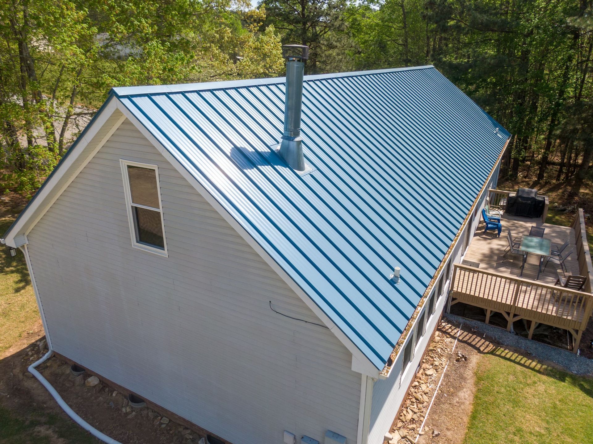 An elevated, angled view of a light-gray house featuring a bright blue standing-seam metal roof and a backyard deck.