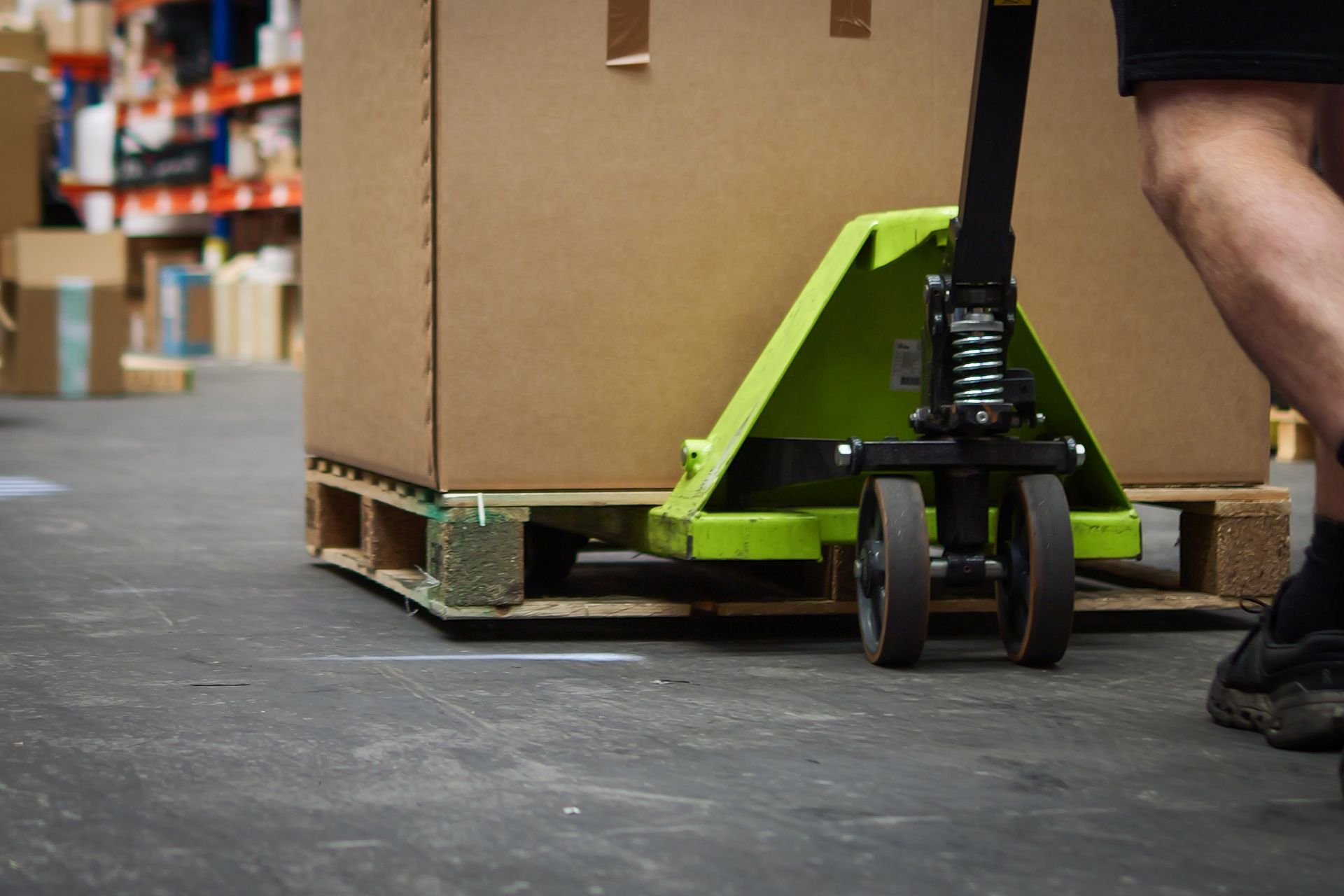 A person using a green pallet jack to move a large cardboard box on a wooden pallet in a warehouse.