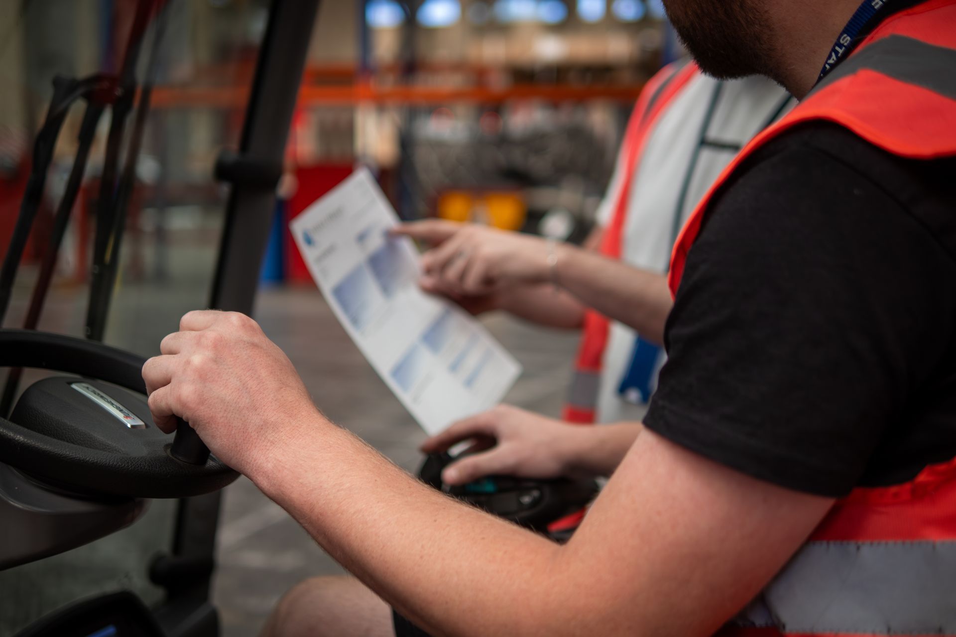 Man driving a forklift, reading paperwork with another person in a warehouse setting.