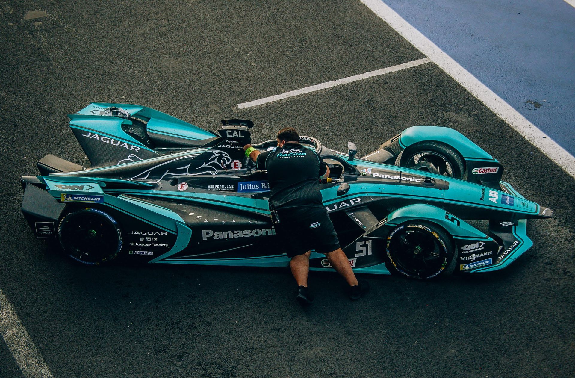 Mechanic working on a turquoise Jaguar Formula E race car in the pit lane.