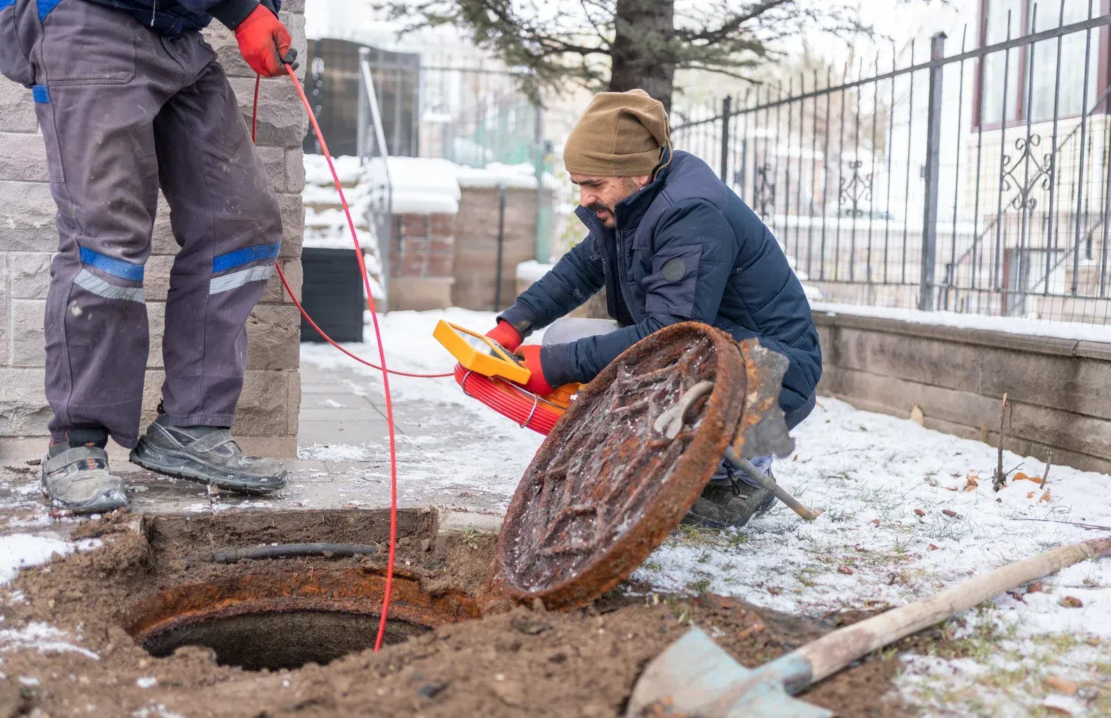 Two workers inspecting a manhole outdoors in snowy conditions using electrical equipment.