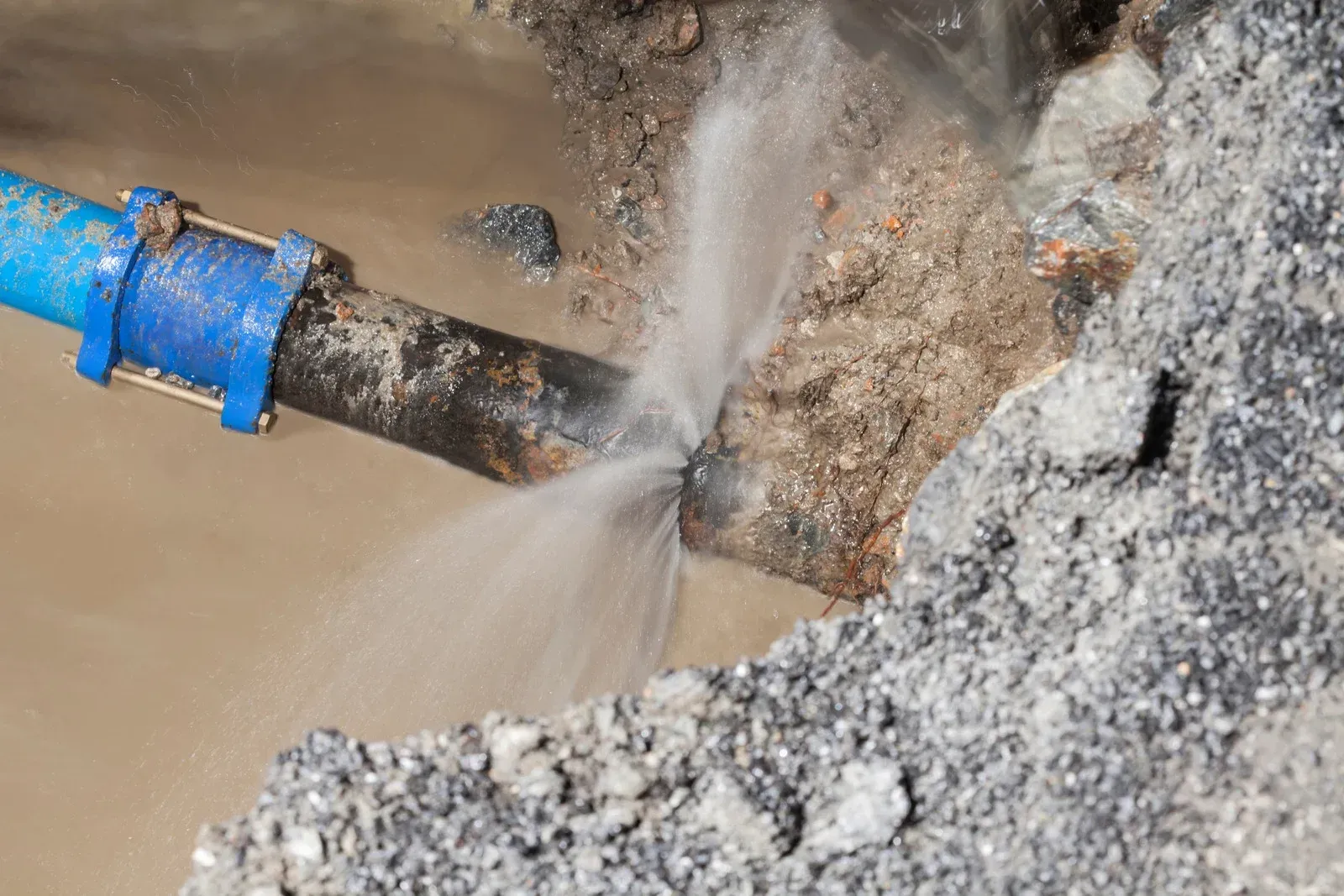 Broken water pipe with water spraying out, in a dirt and gravel setting.