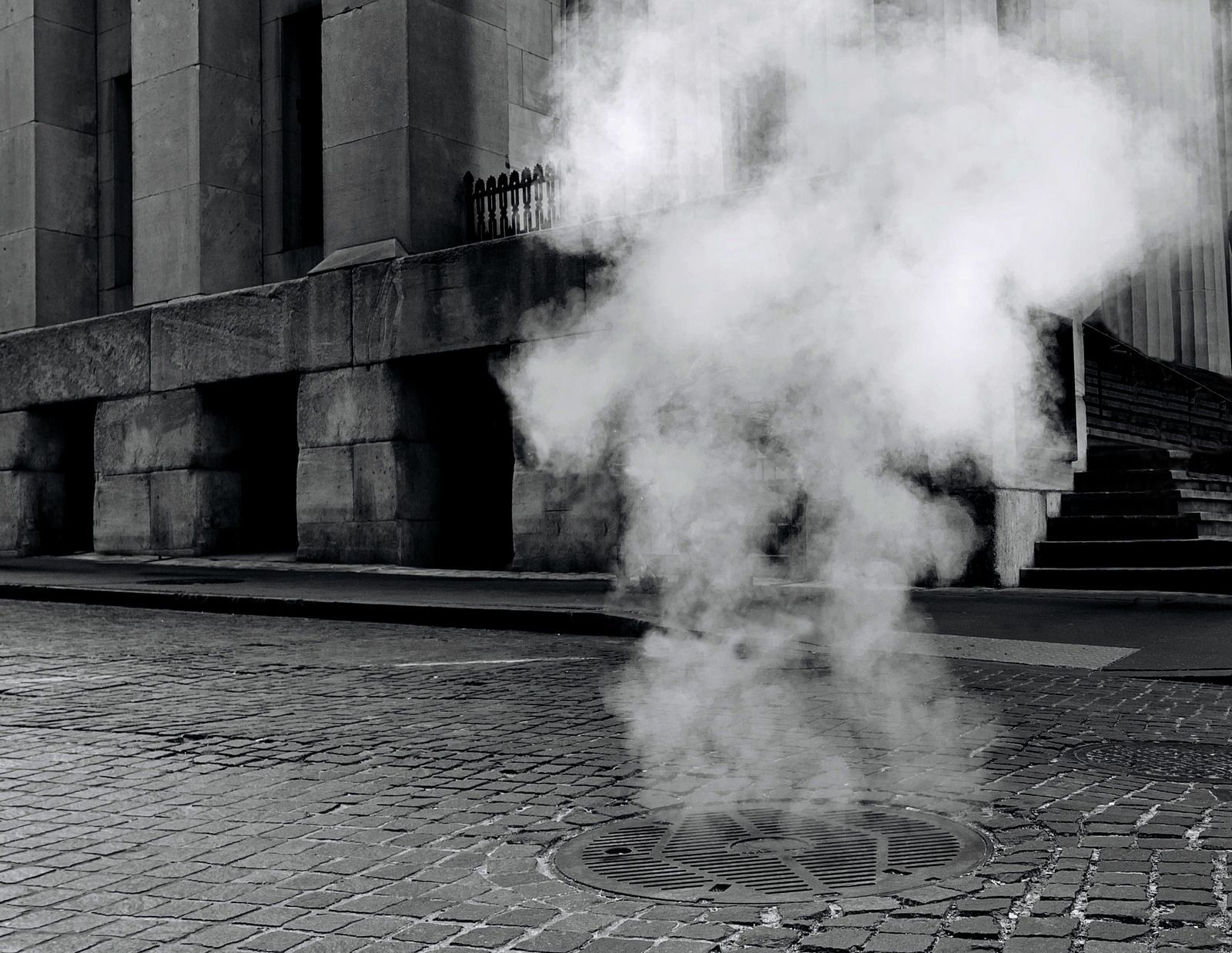 Steam rising from a manhole cover in a cobblestone street near a building.