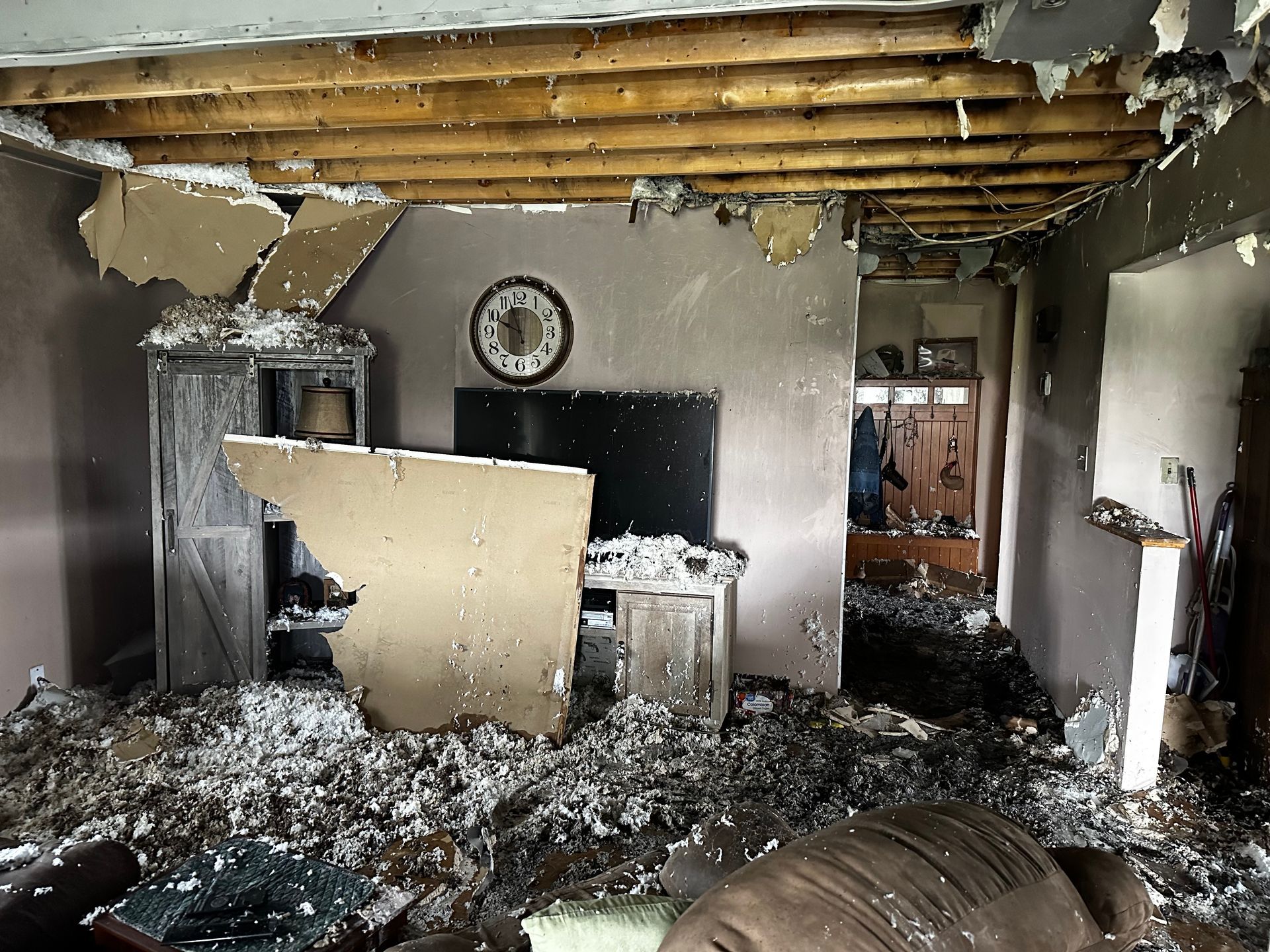 Interior room damaged by fire; debris on floor, ceiling exposed, clock on wall, charred furniture.