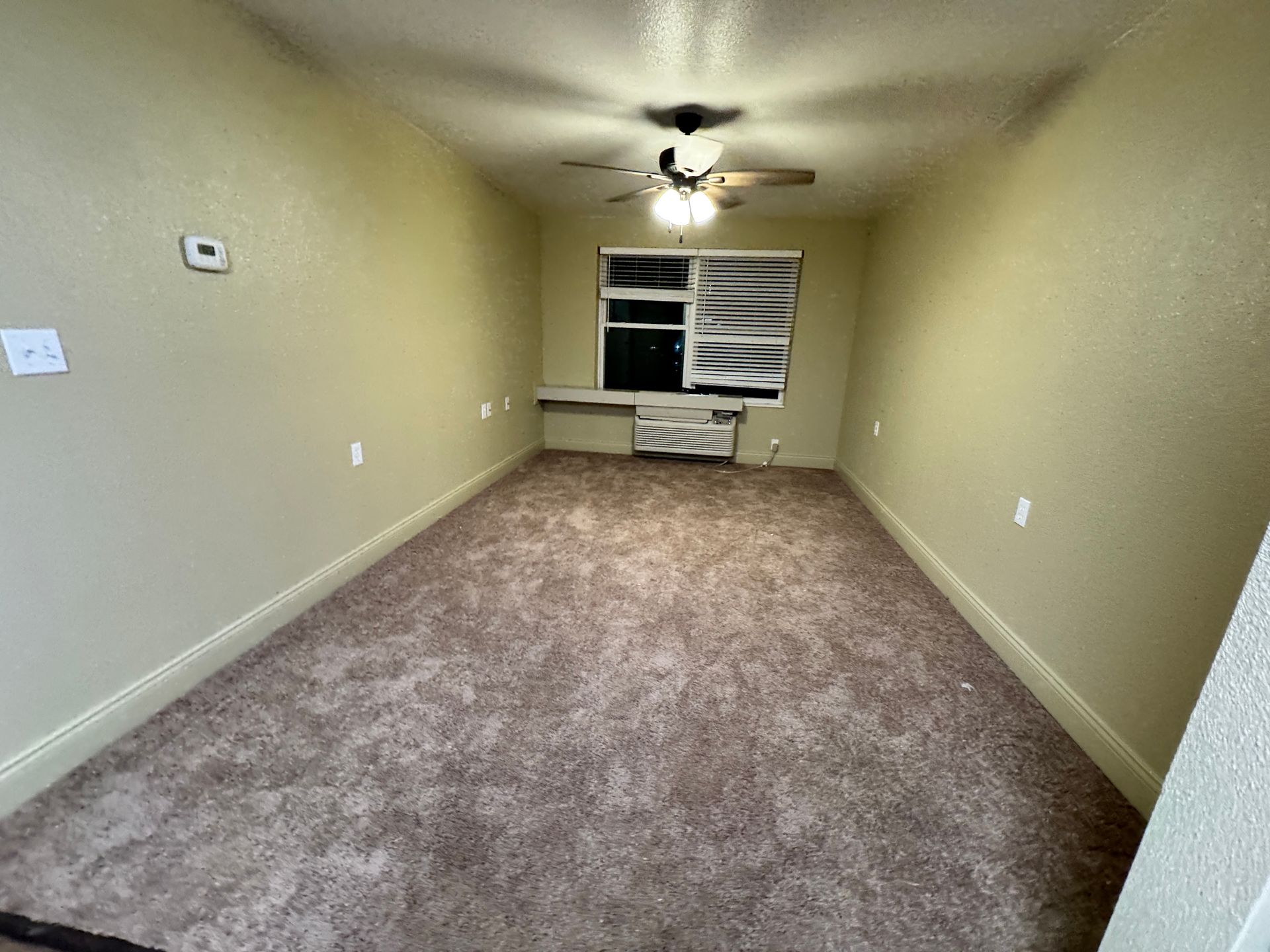 Empty room with tan walls, brown carpet, window with covered blinds, and ceiling fan.