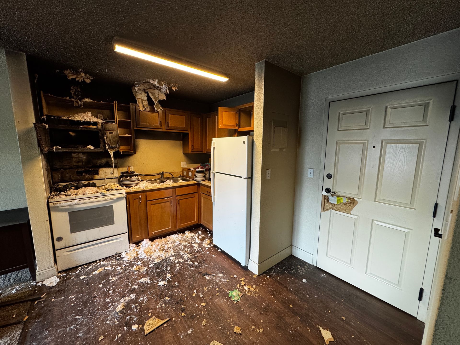Kitchen severely damaged by fire; cabinets, appliances, ceiling, and floor show heavy charring. Debris scattered.