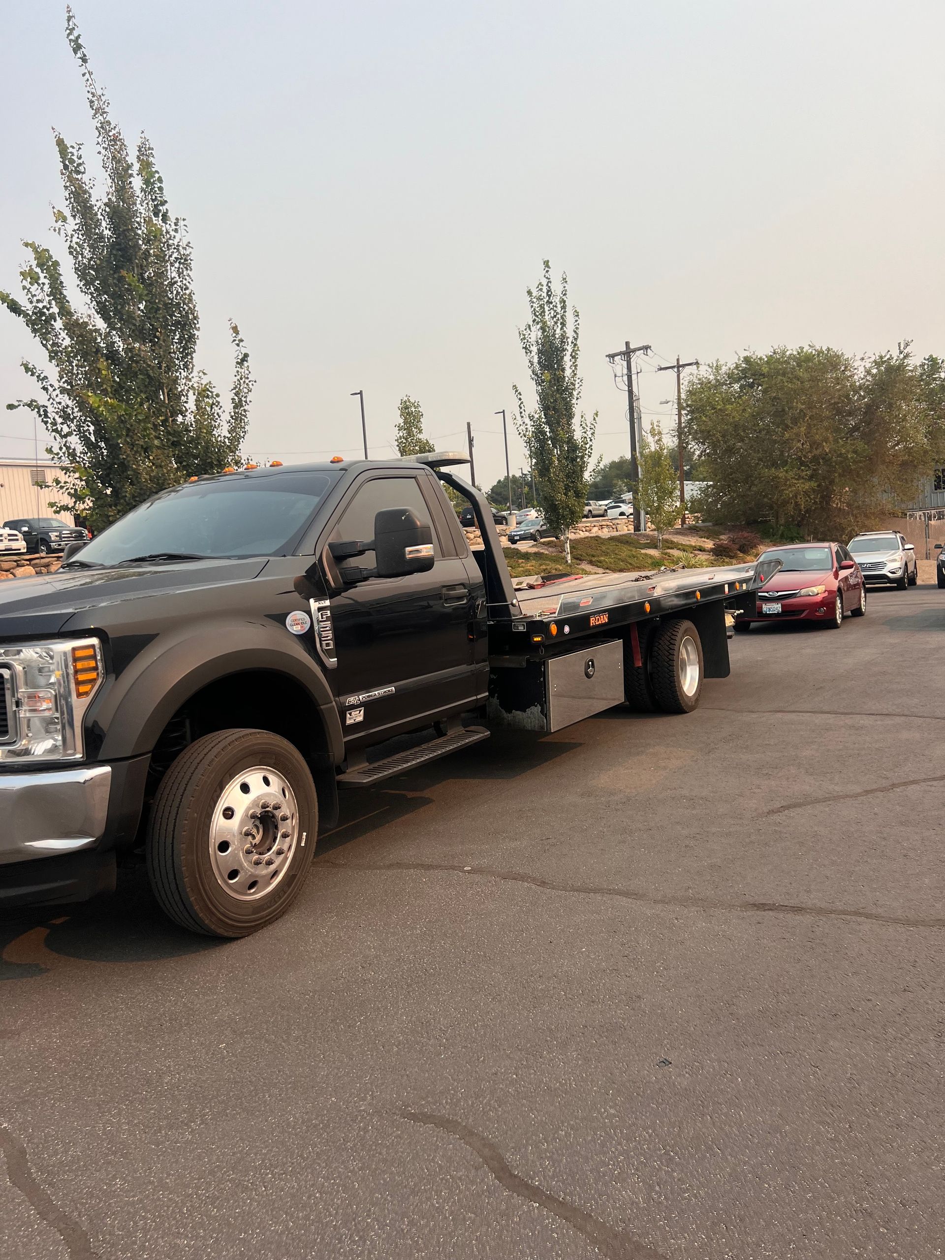 Black flatbed tow truck parked on a road near trees, with cars lined up behind it at dusk