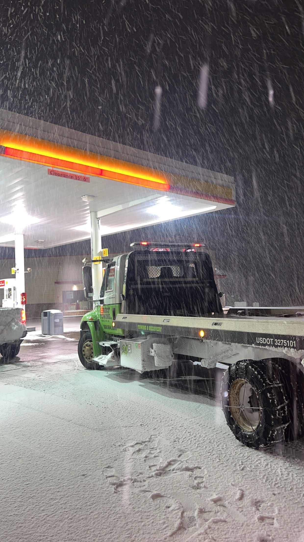 Tow truck at a snowy gas station at night under bright canopy lights, with snow falling heavily.