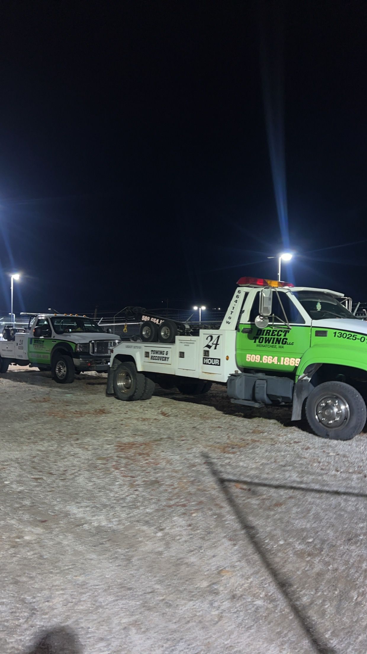 Green tow truck and other utility trucks parked on a gravel lot at night under bright lights.