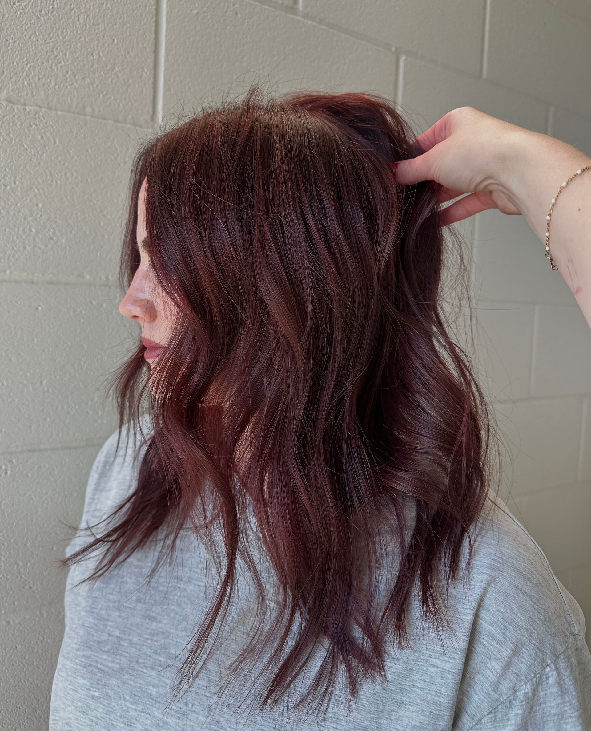 Woman with wavy, dark red hair, being styled with a hand in the hair; white brick wall in the background.