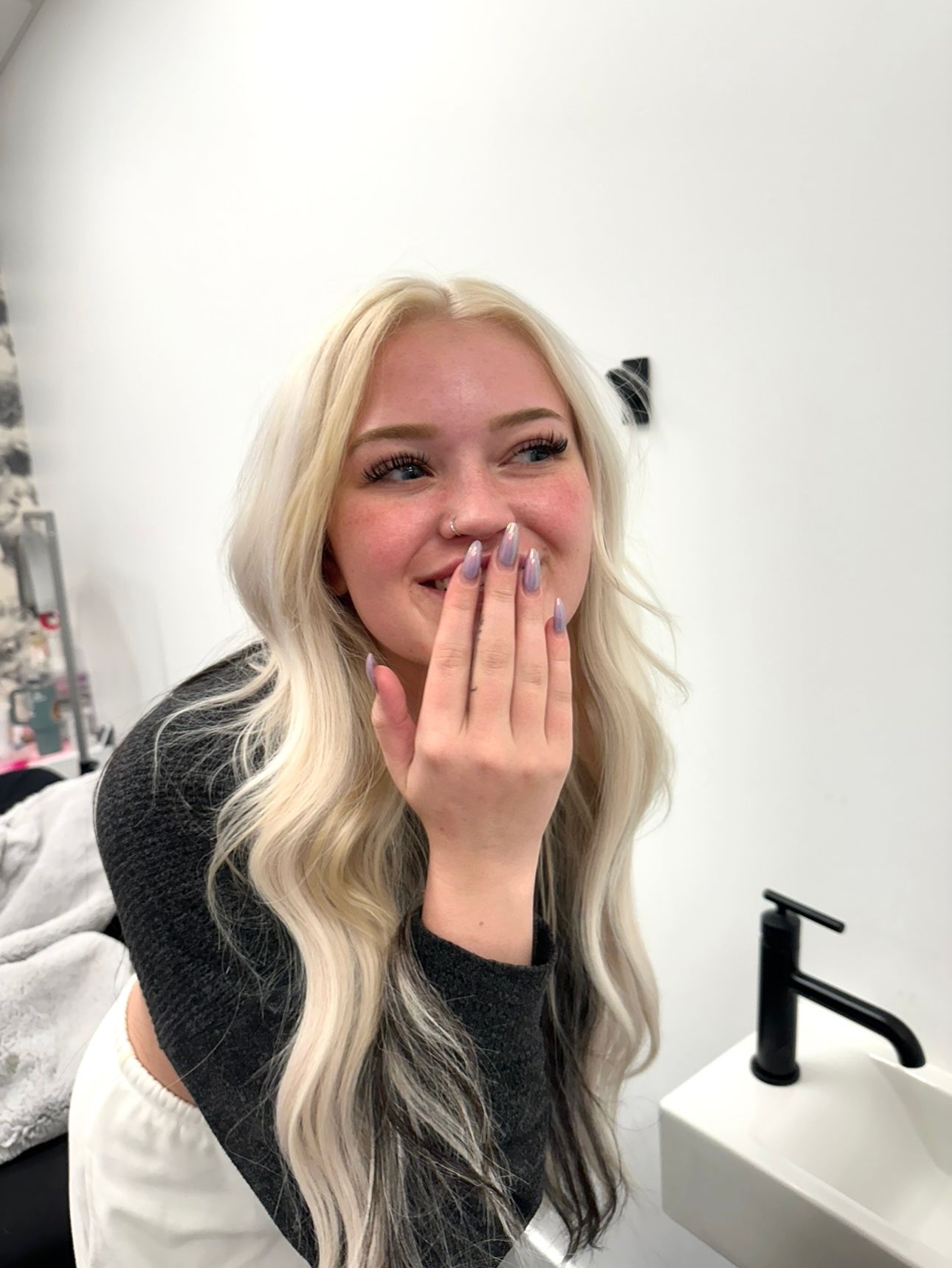 A woman with blonde hair is covering her mouth with her hand while looking lash extensions