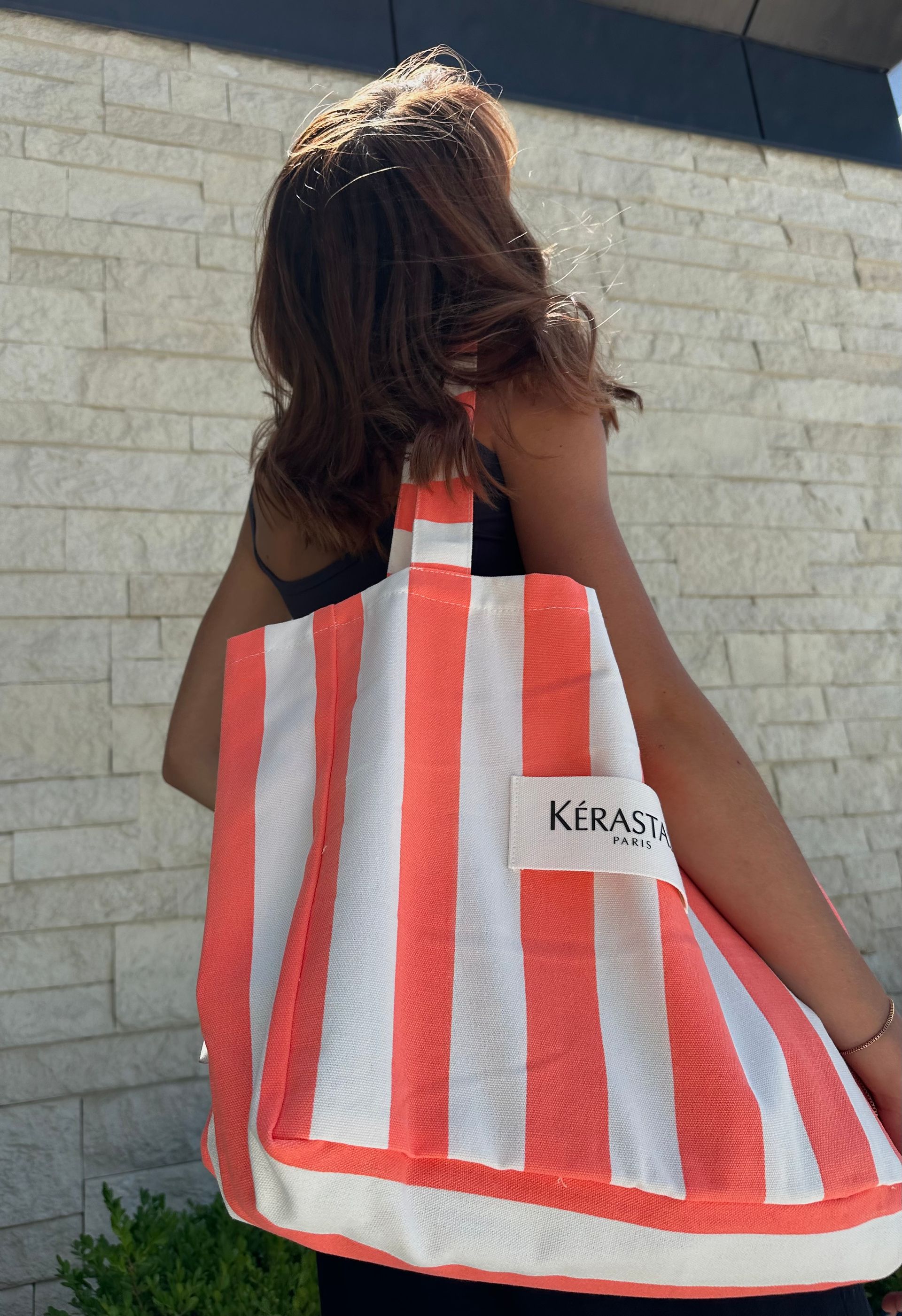 Woman carrying a coral and white striped tote bag, standing in front of a brick wall.