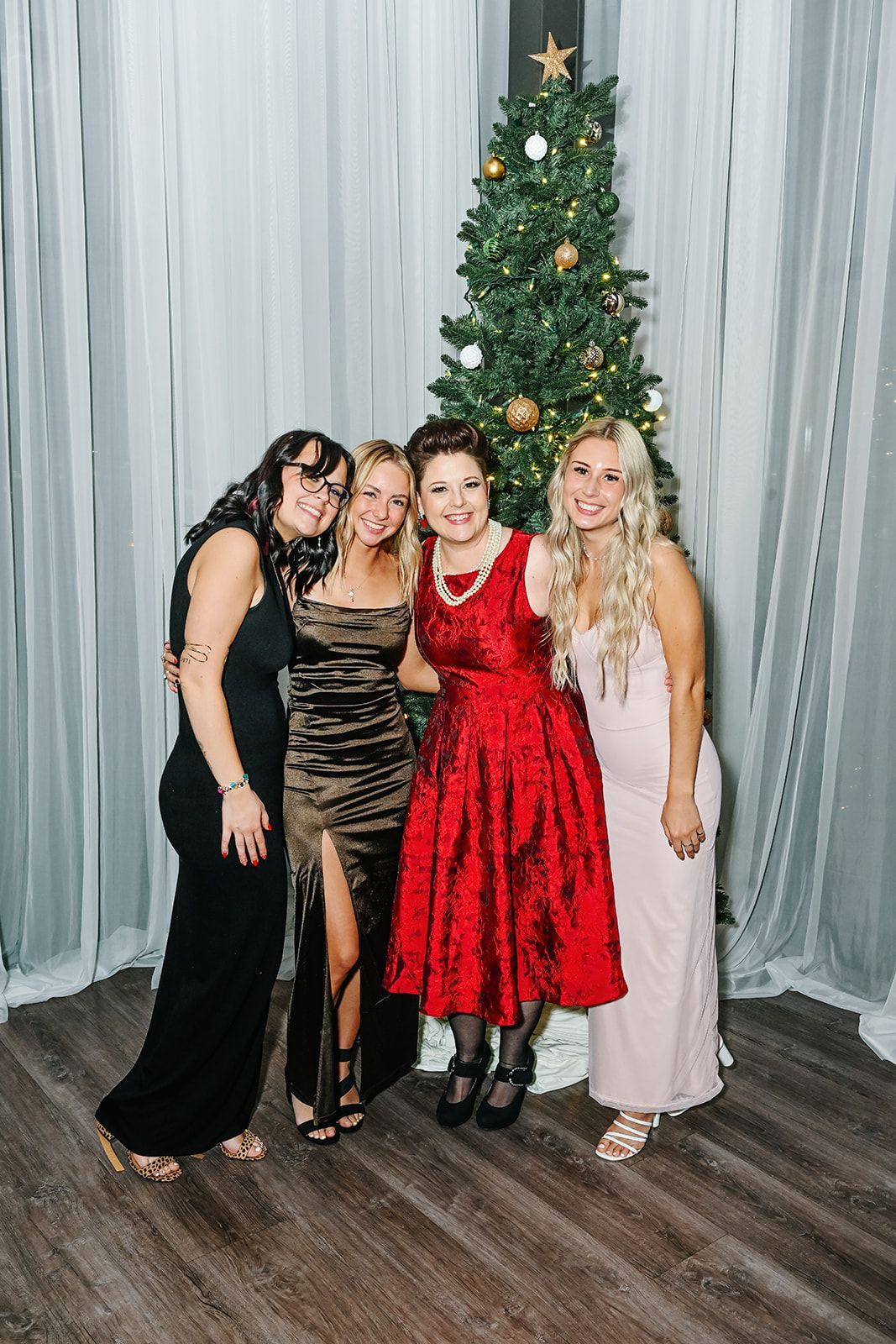 Four people in formal wear pose by a Christmas tree; they smile and embrace in front of a white curtain.
