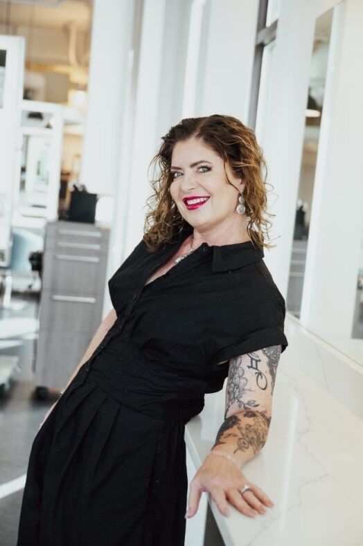 Sarah, director of Guest Experience at Ottalaus Salon, wearing glasses and a black dress is sitting in a blue chair.