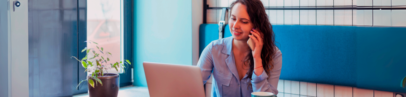 Mulher ao telefone ao lado de um laptop em um escritório moderno com cadeiras azuis e uma planta.
