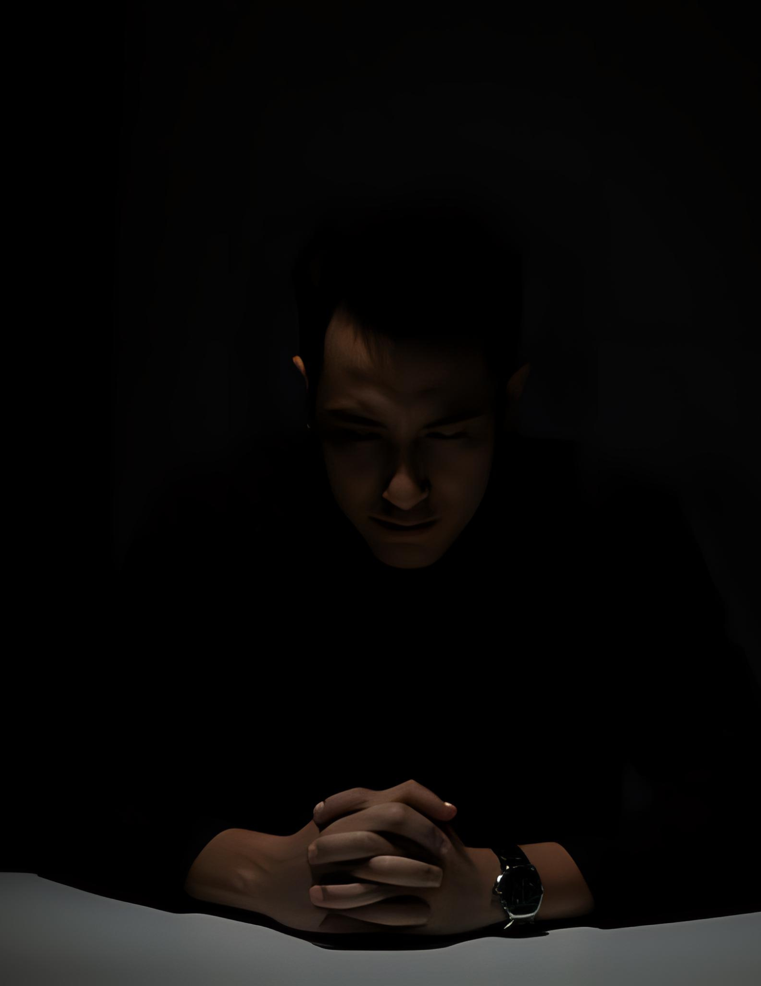 Man in dark room, hands clasped on table.  Face is lit, showing a concerned expression.