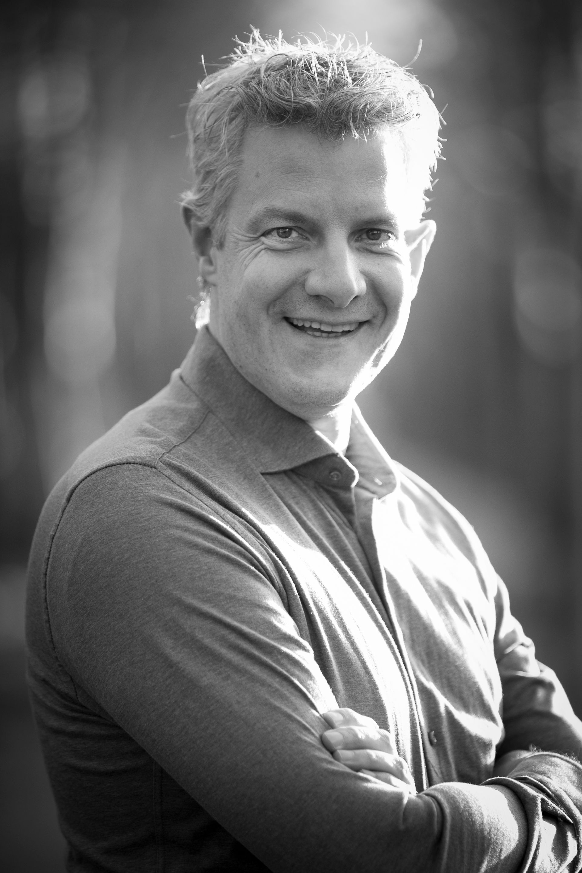 Man with graying hair smiling, arms crossed, outdoors, soft focus background.