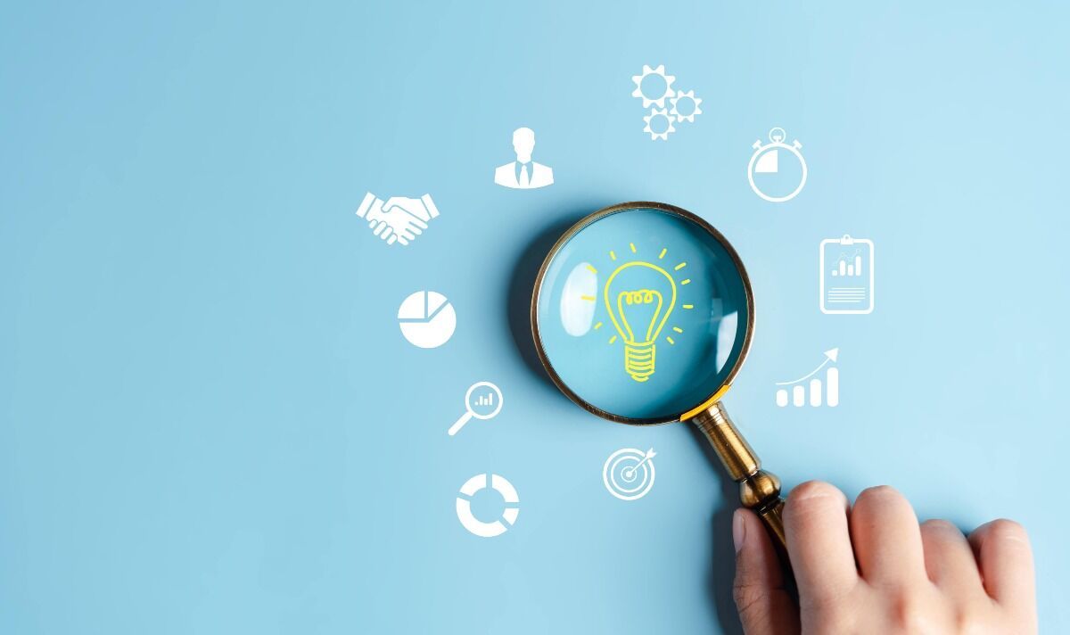 Hand holding magnifying glass over a lightbulb icon, surrounded by business-related icons, all on a blue background.