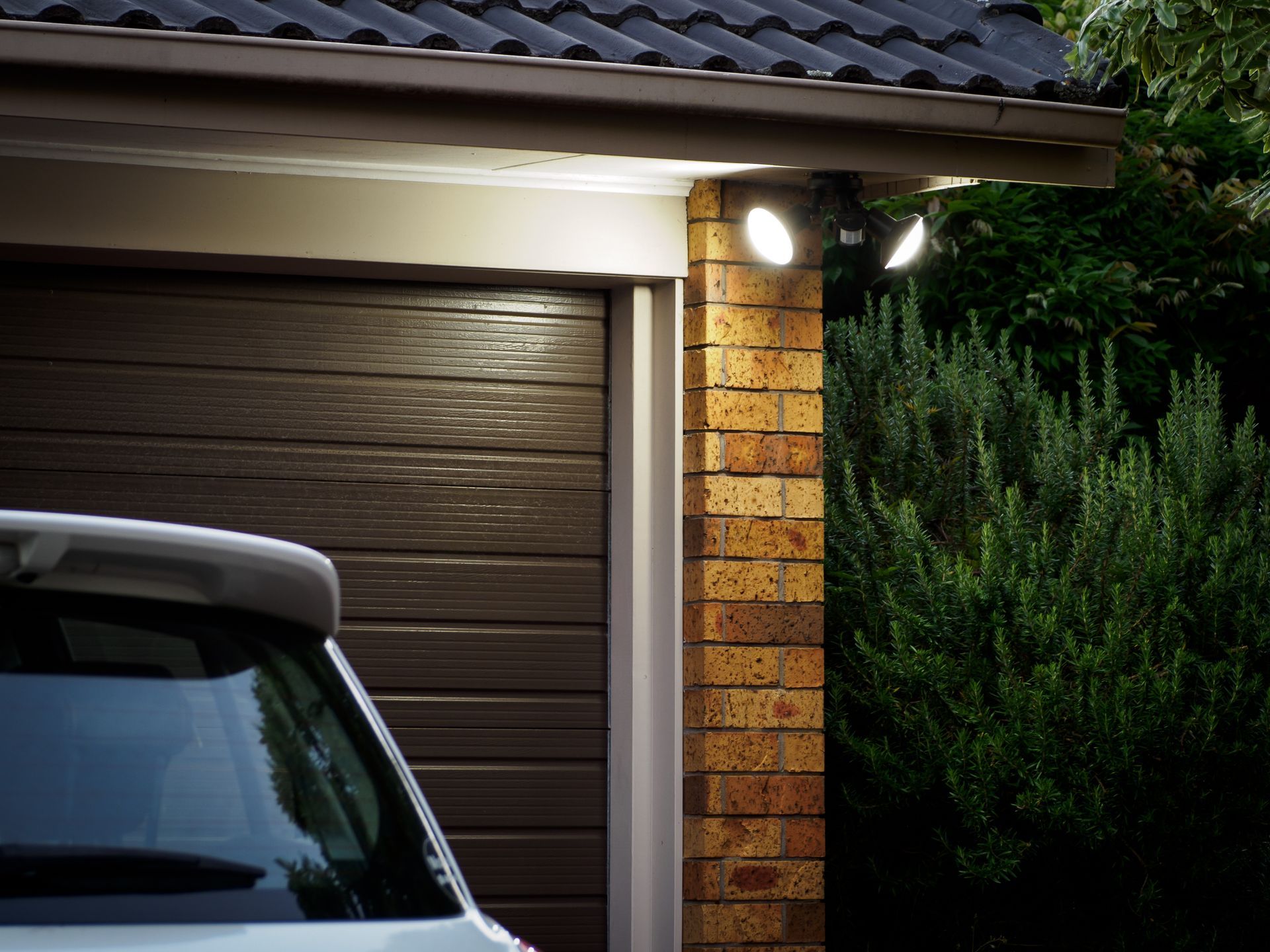 Security lights illuminate a garage door and a brick pillar next to a green bush and a car — Ryphen Electrical in Highton, VIC