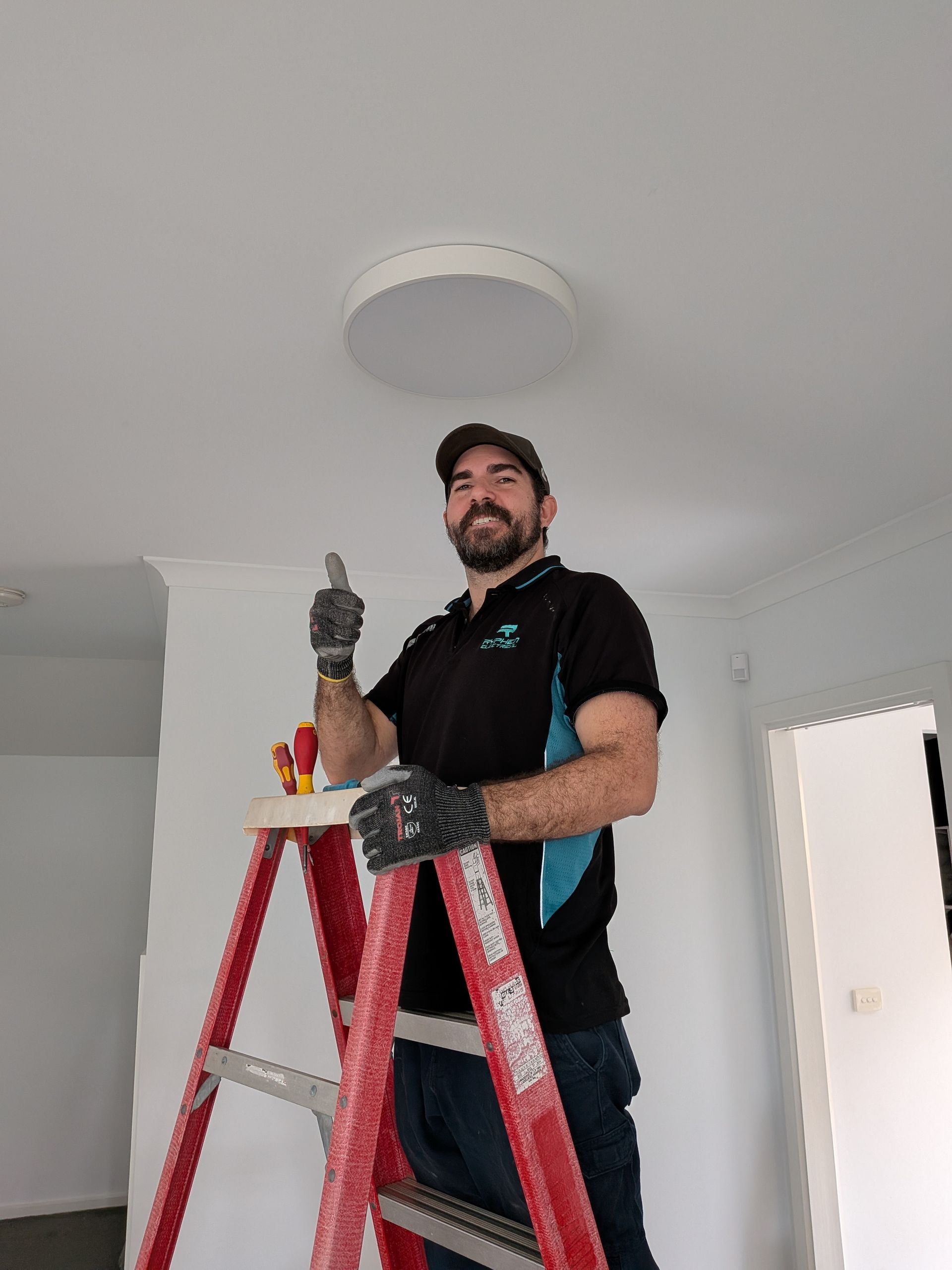 Electrician on a ladder installing a ceiling light, giving a thumbs up.