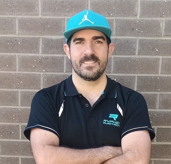 Man wearing a teal cap and black polo shirt with arms crossed, in front of a brick wall — Ryphen Electrical in Highton, VIC