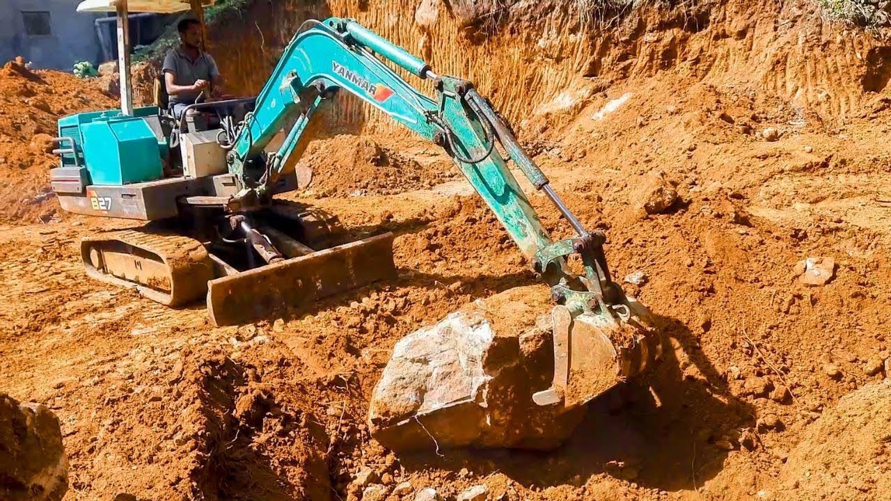 Turquoise excavator digging in a muddy earth excavation site with a large rock in the bucket