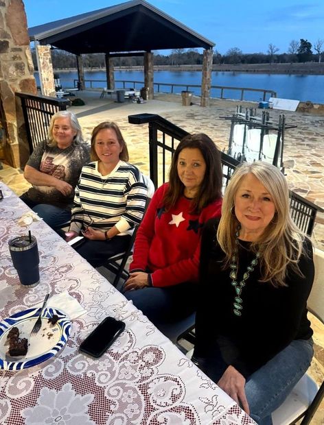Four people seated at an outdoor lakeside table, smiling beside a stone patio and pavilion.
