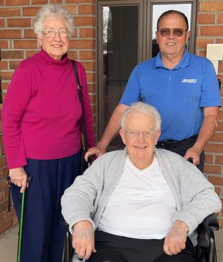 An elderly woman in a wheelchair is surrounded by two other people