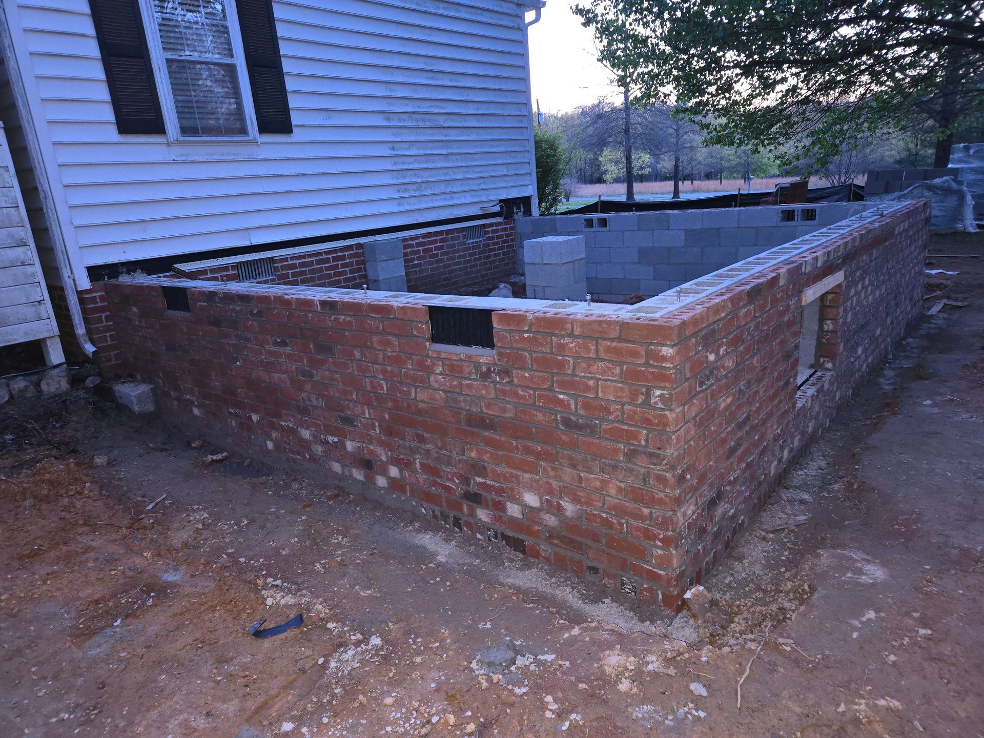 A brick foundation wall under construction attached to the side of a white-sided house on an dirt lot.