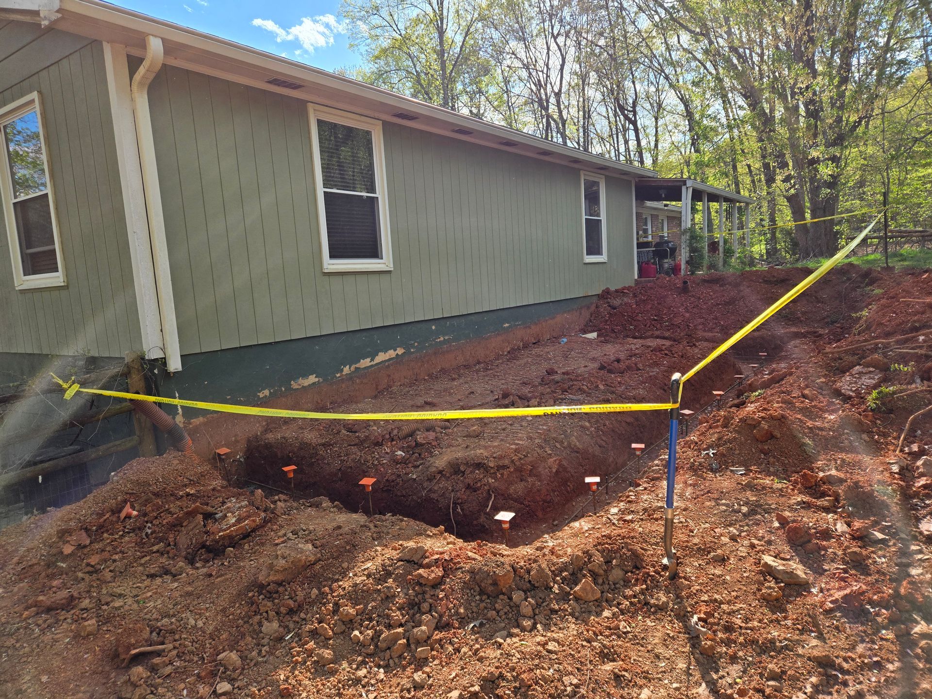 A side view of a green house with a dug-out trench marked by orange flags and yellow caution tape in the front yard.