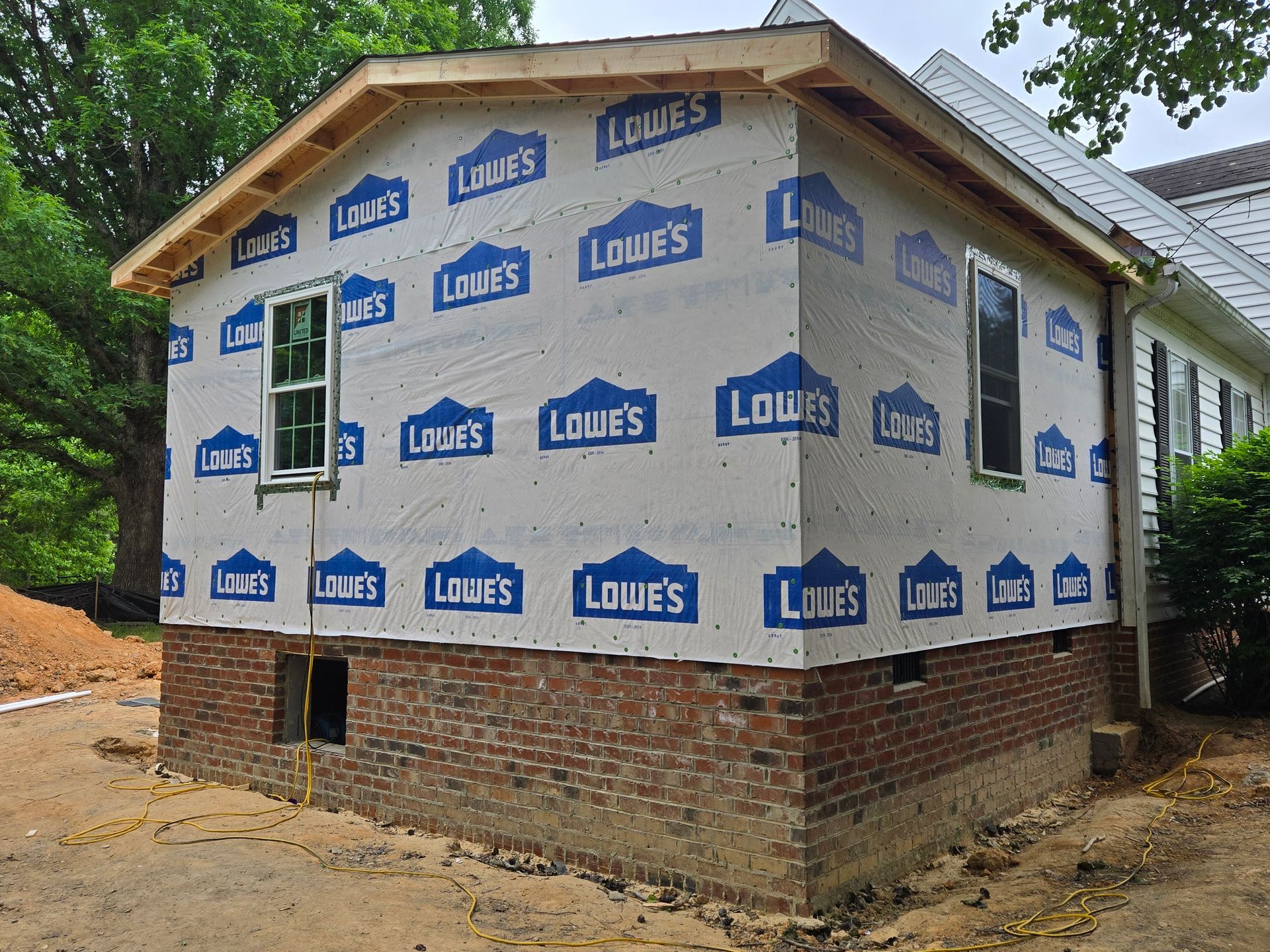 The side of a house under renovation, featuring exposed brick foundation, new framing, and white Lowe's house wrap.