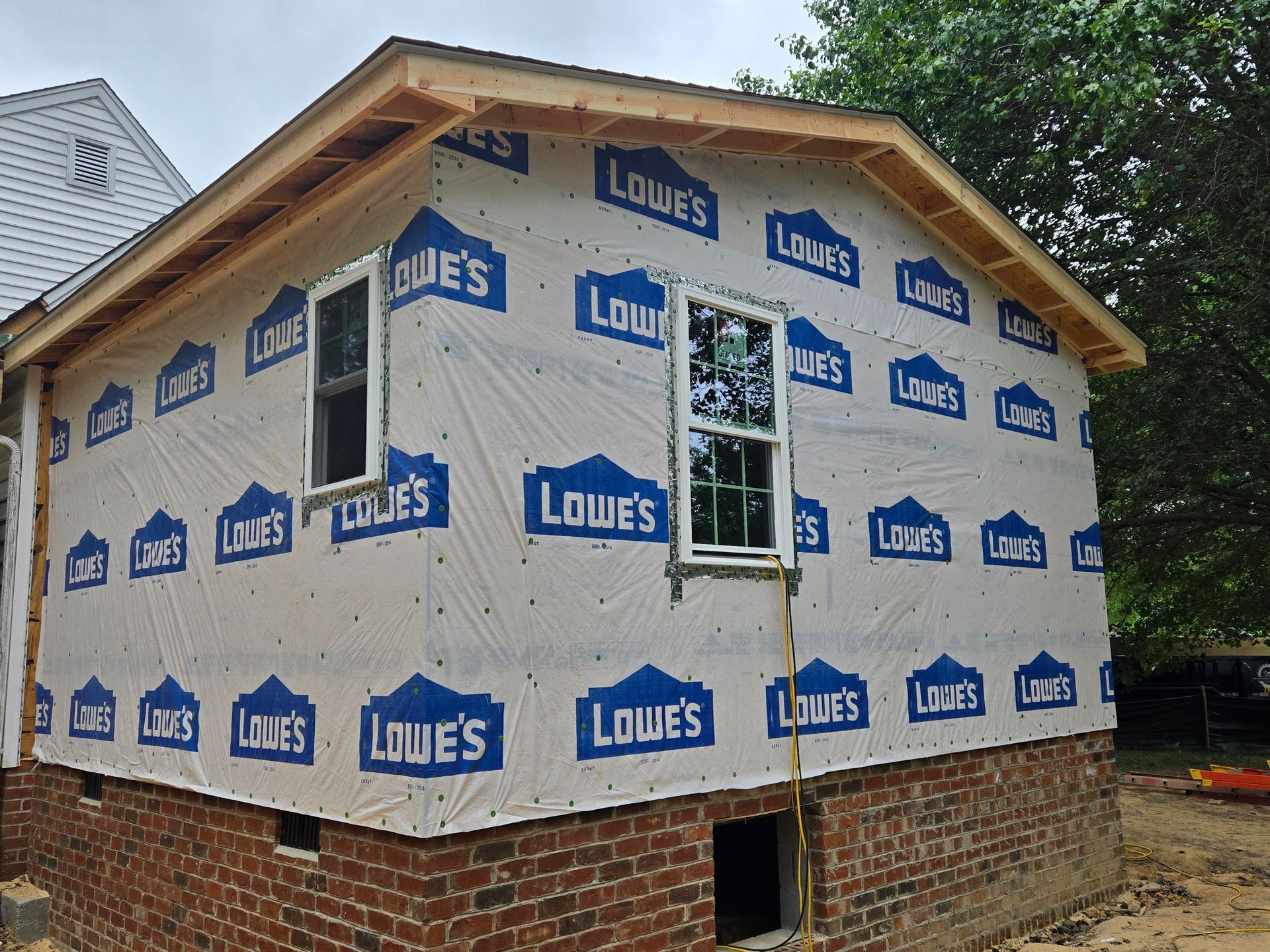 A residential home addition under construction with white house wrap featuring Lowe’s logos over a brick foundation.